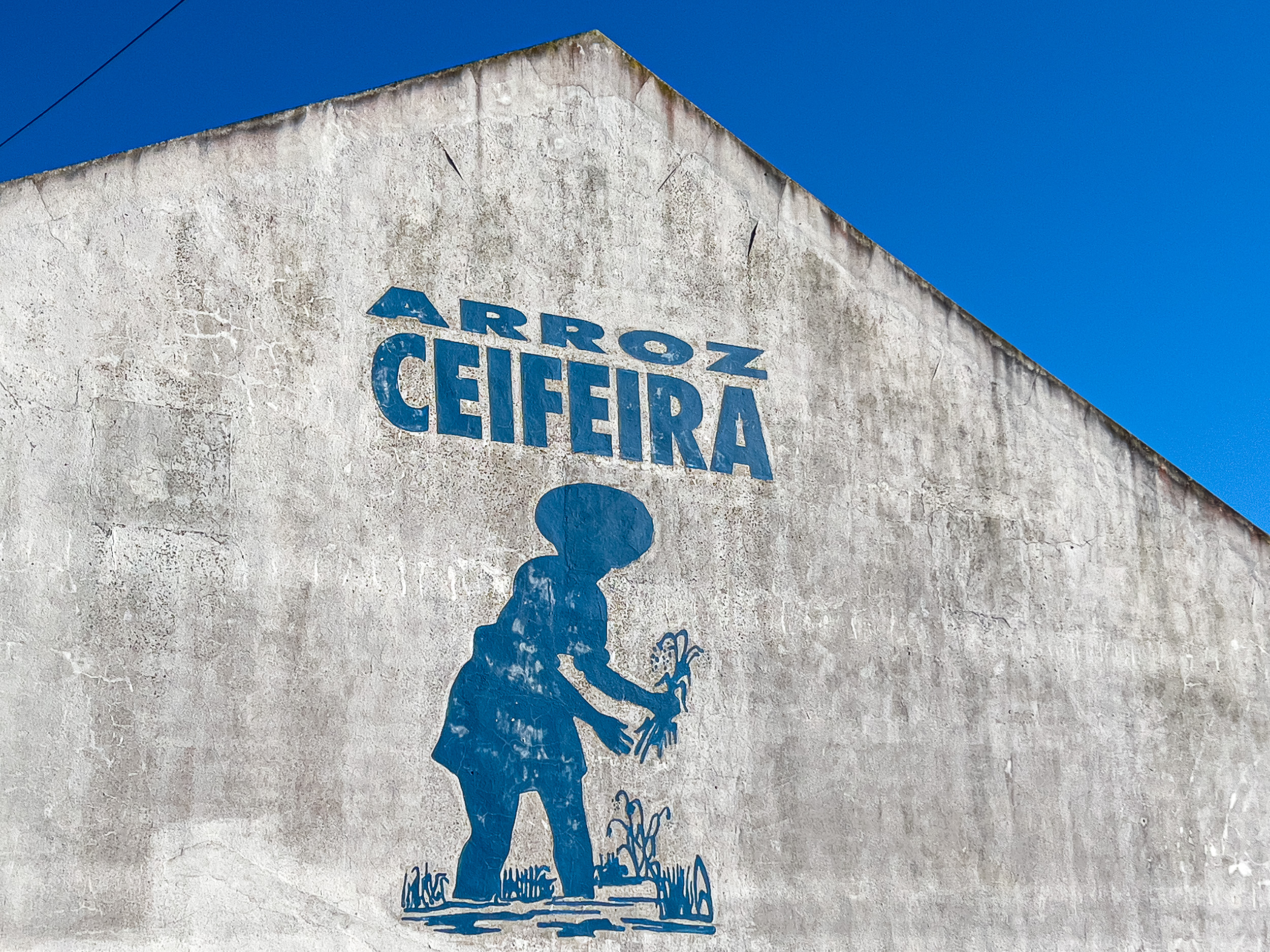A whitewashed facade with the classic blue mural 'Arroz Ceifeira' depicting a rice harvester