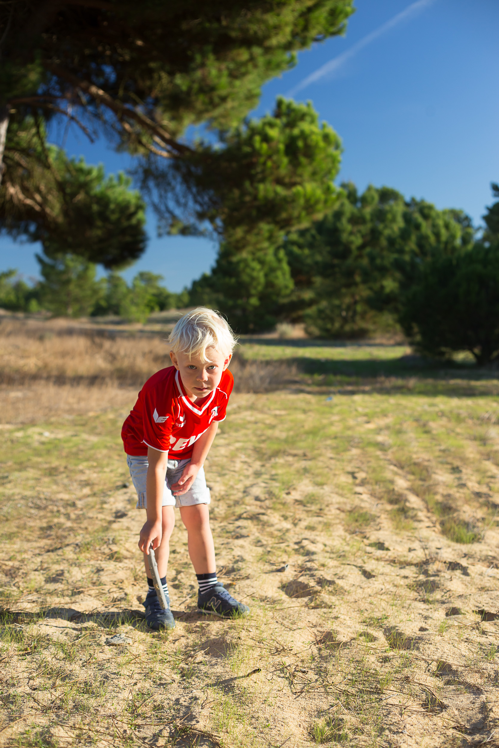 Un garçon blond en maillot rouge jouant avec un bâton dans le sol sablonneux d'une forêt de pins par une journée ensoleillée.