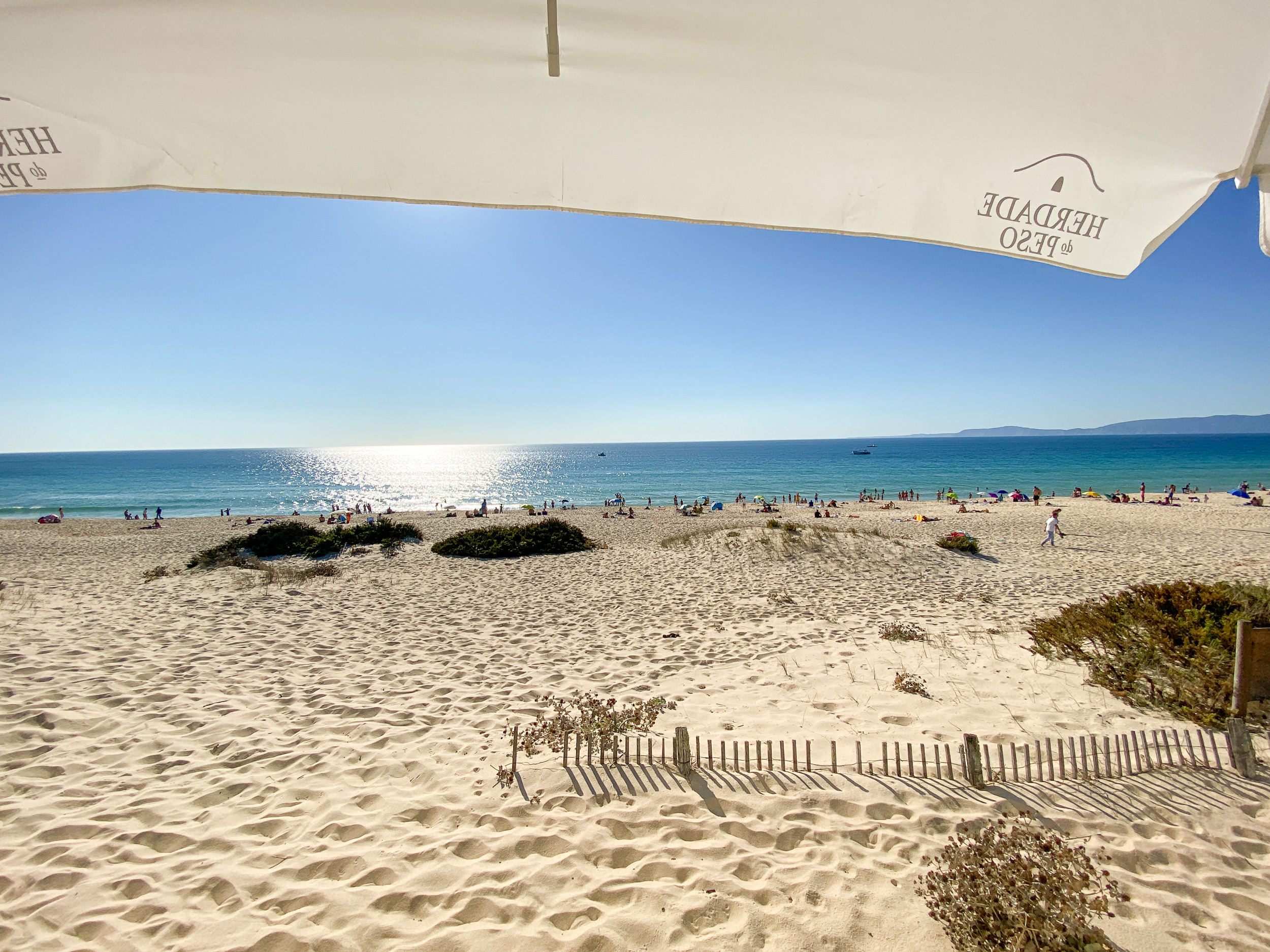 Vue sous un parasol blanc de la grande plage de sable Praia da Comporta et de la mer scintillante.
