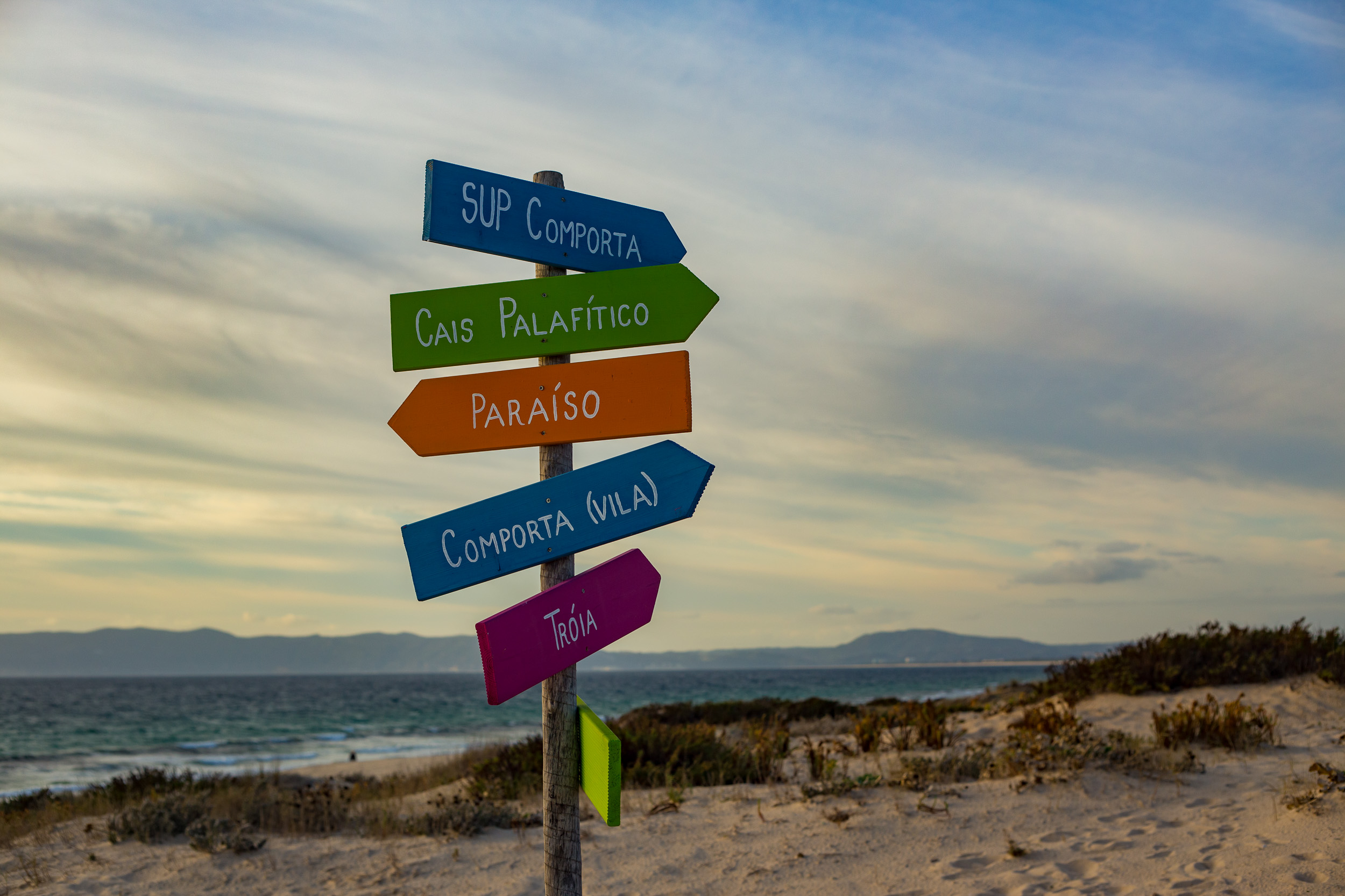 Un panneau indicateur avec des panneaux en bois colorés sur la plage de Comporta, pointant dans différentes directions.