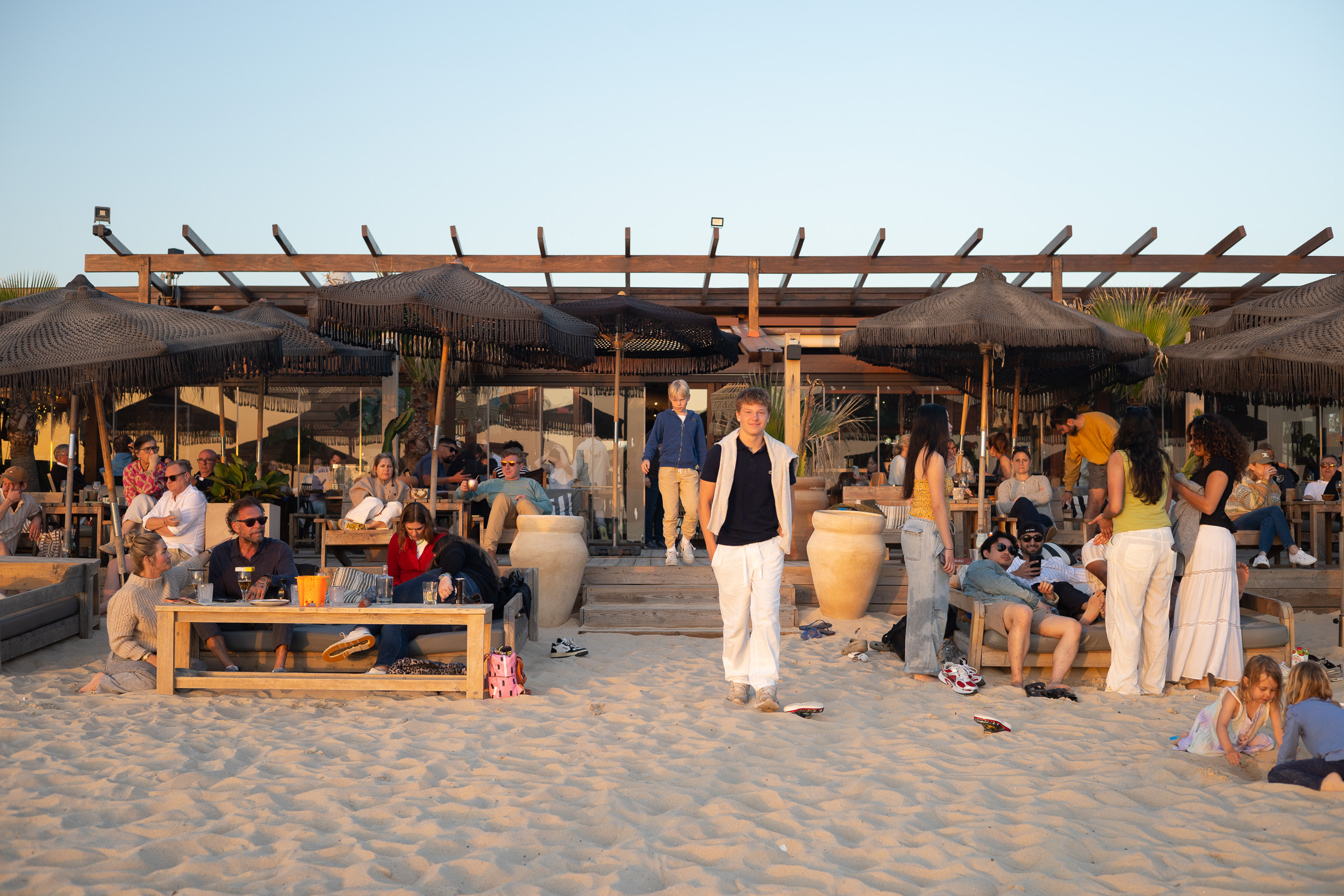 Des clients se détendant sur la terrasse en sable du Comporta Café sous des parasols sombres dans la lumière de fin d'après-midi.