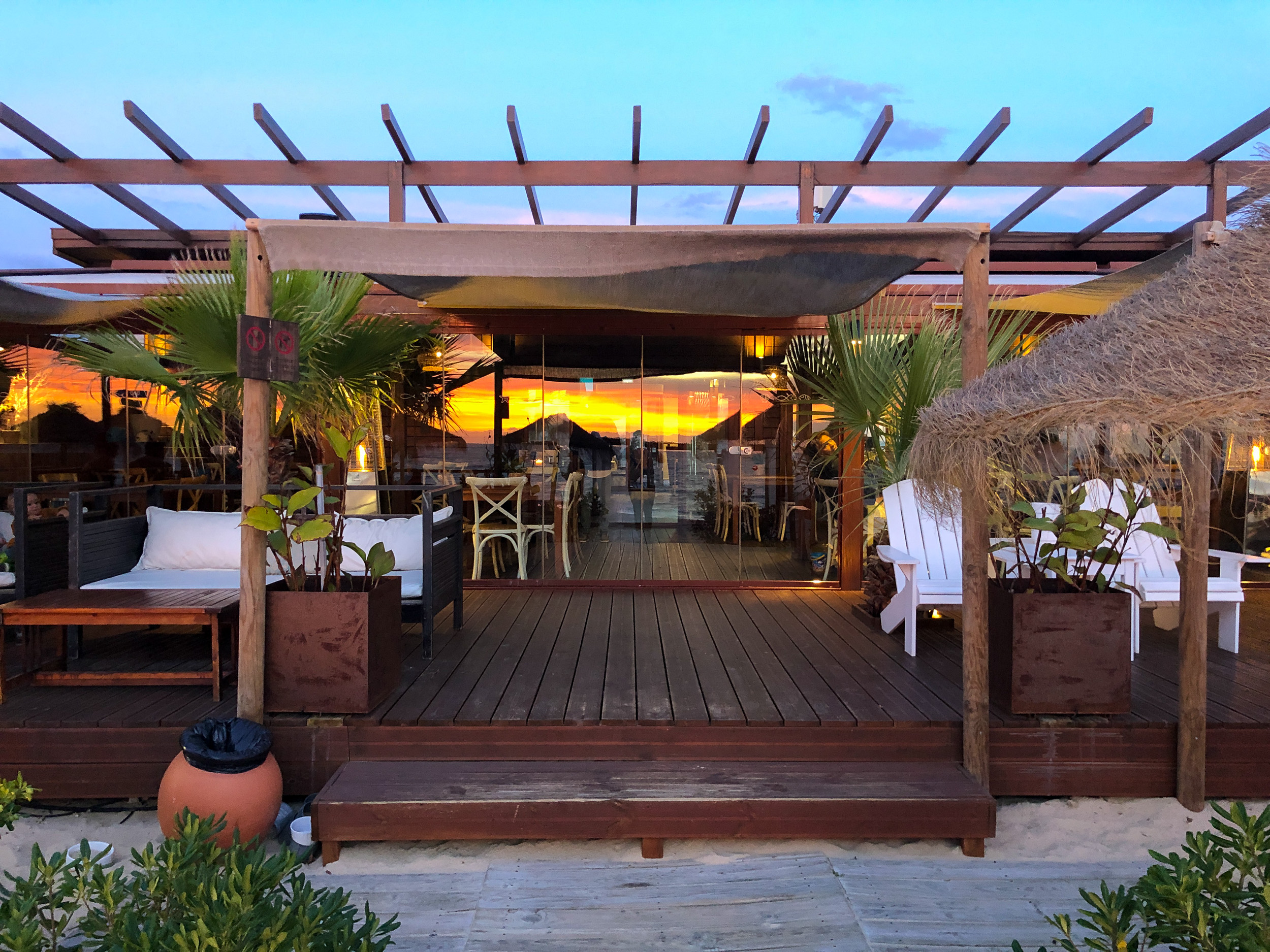 Le coucher de soleil éclatant se reflète dans la façade en verre sur la terrasse en bois du Comporta Café.