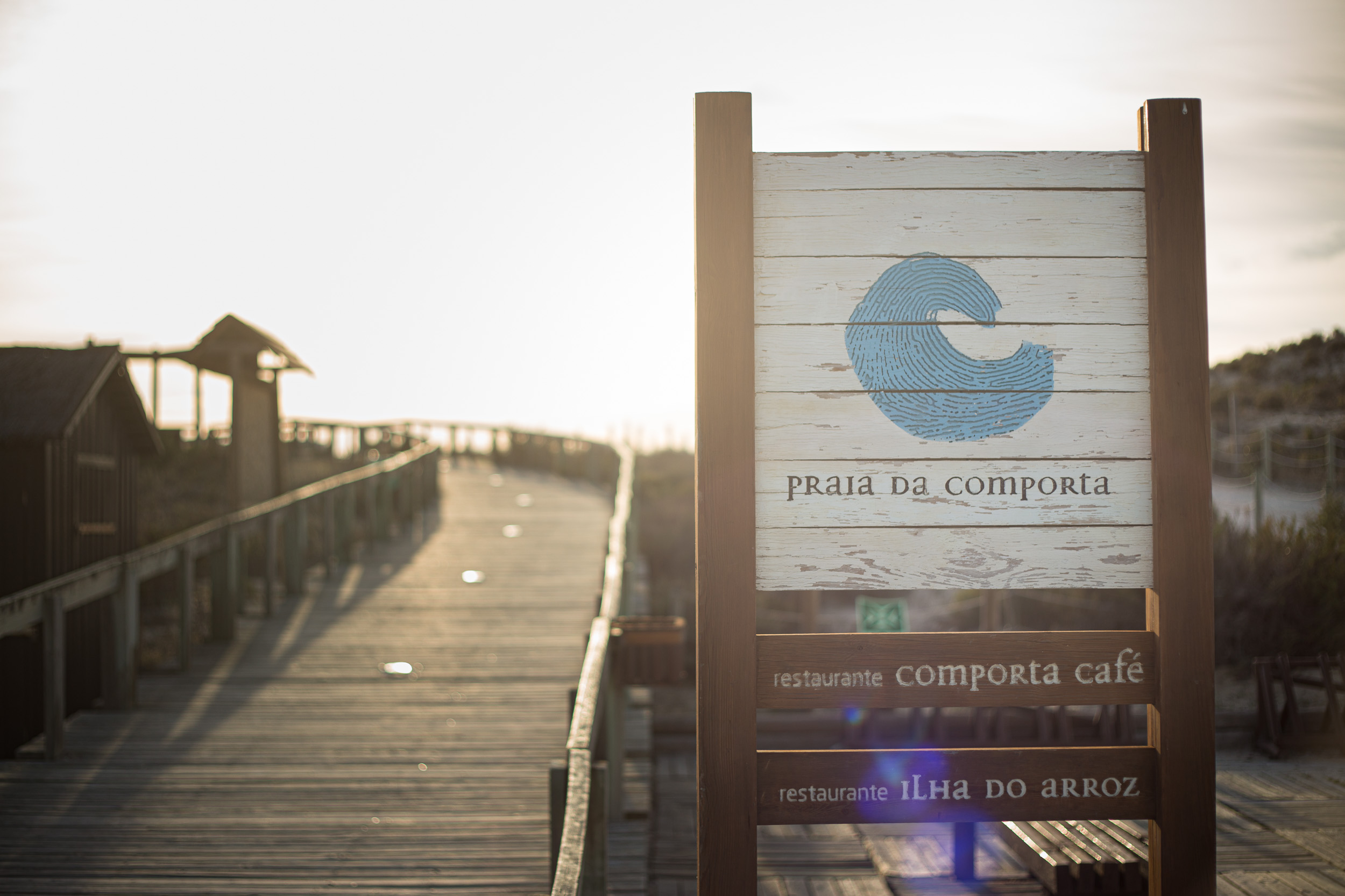 Ein Holzschild am Holzbohlenweg zum Praia da Comporta mit den Wegweisern zum Comporta Café und Ilha do Arroz im goldenen Gegenlicht.