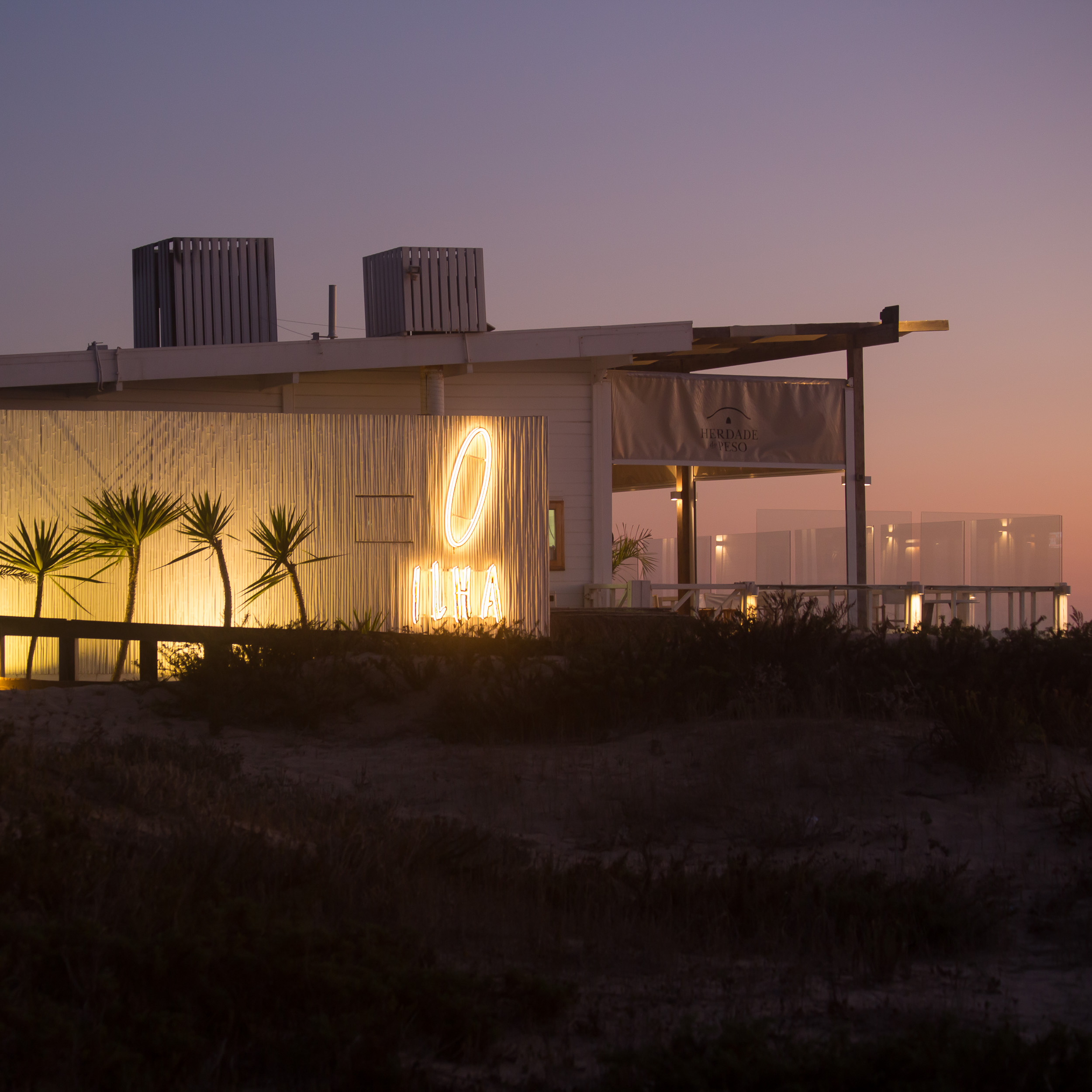 Das leuchtende ILHA-Neonschild an der weißen Lamellenfassade des Ilha do Arroz Restaurants vor lila-rosa Abendhimmel.