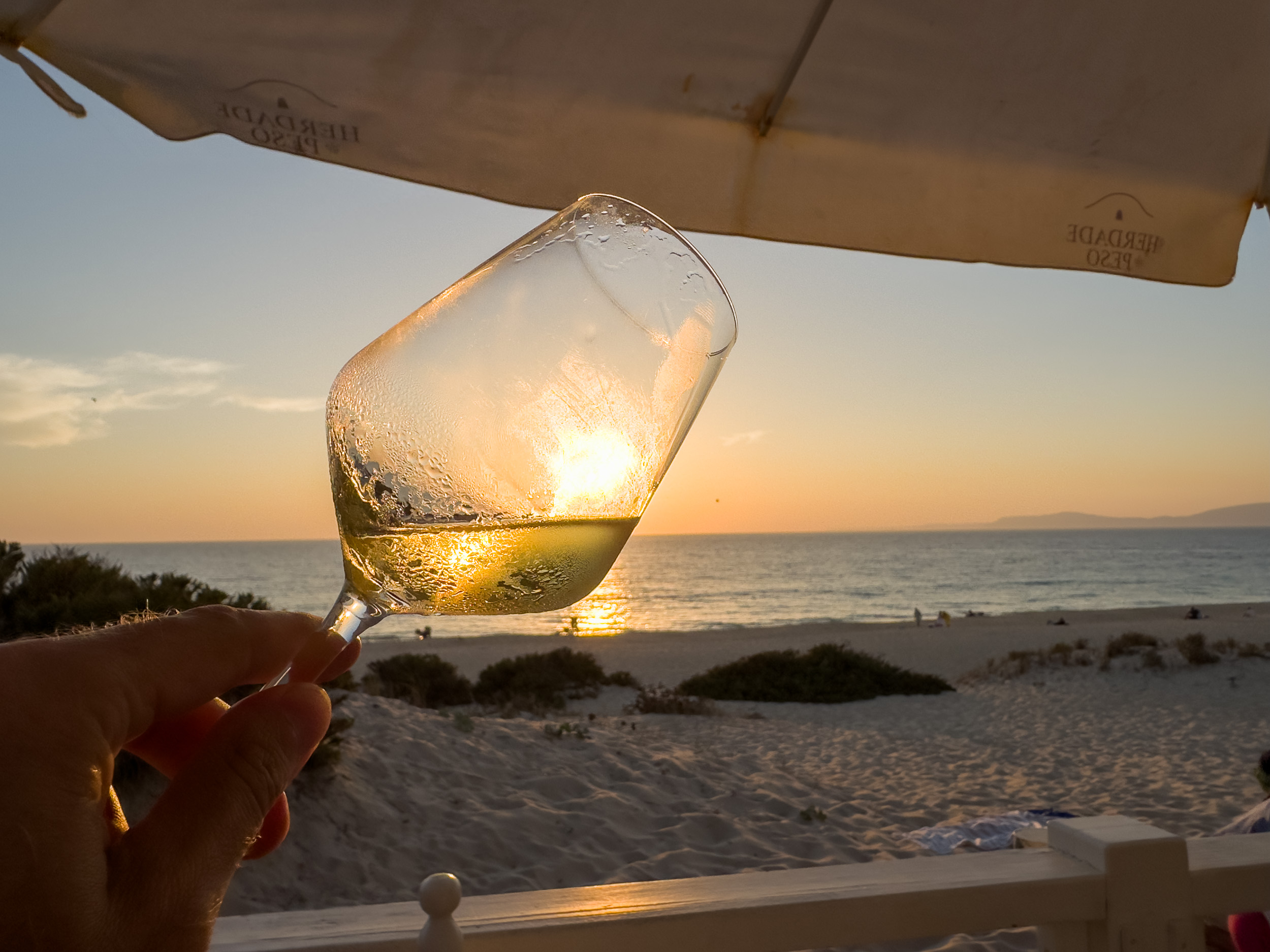Eine Hand hält ein kühles Glas Weißwein im Gegenlicht der untergehenden Sonne, dahinter der weiße Sandstrand und das Meer.