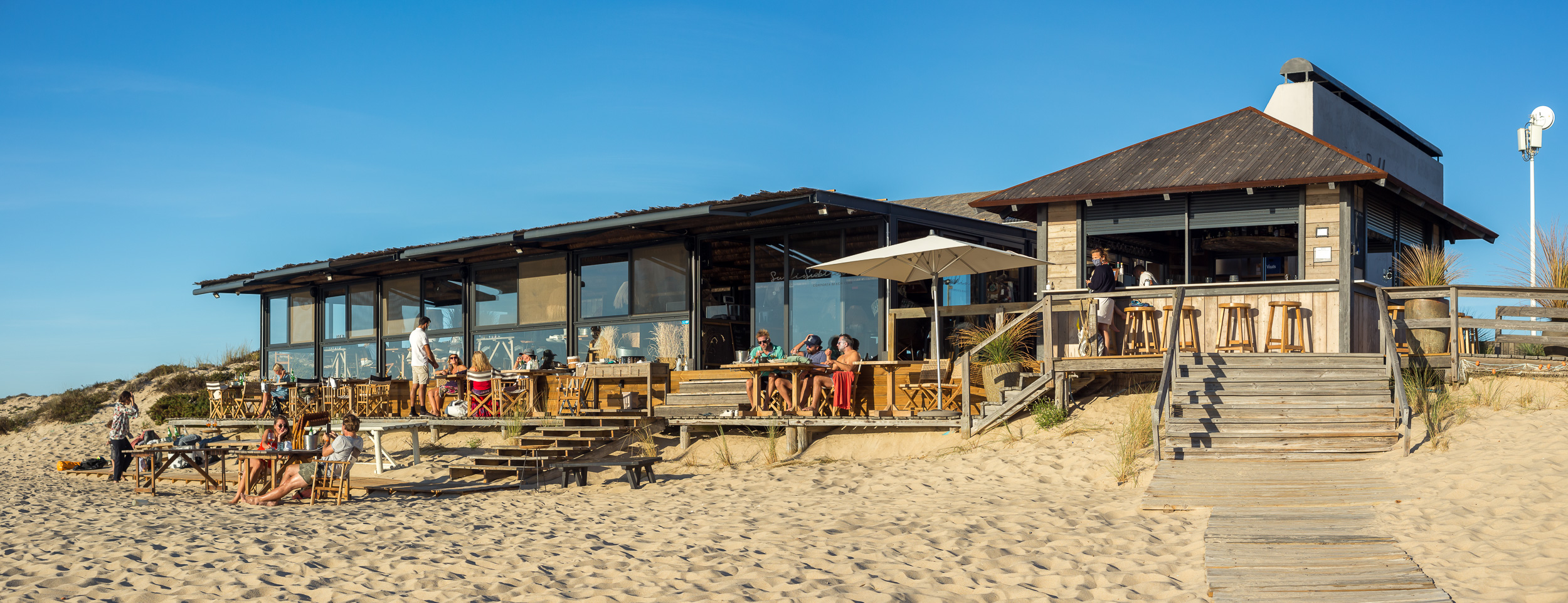 Breite Panorama-Außenansicht des Holzgebäudes vom Sublime Beach Club mit Außenterrasse direkt am Sandstrand von Comporta.